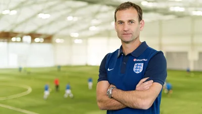 Dan Ashworth in an England training top, arms folded, on an indoor pitch.
