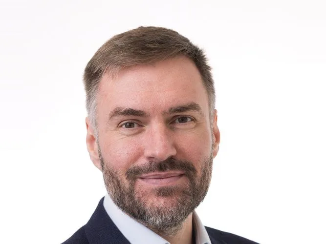 Andrew Hulbert, in a navy jacket and open-collared shirt, against a white studio background.